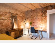 Zimmer mit unverputzter Ziegelwand und Gewölbedecke. Einrichtung umfasst einen Holztisch mit Metallbeinen, einen Stuhl, eine Stehlampe, eine Kommode mit Spiegel sowie eine Topfpflanze und eine Tischleuchte.