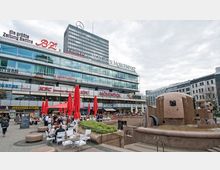 Platz mit mehrstöckigem Gebäude im Hintergrund, das Glasfassaden und Werbeschilder verschiedener Geschäfte und Restaurants zeigt. Im Vordergrund ein Brunnen aus Stein und Sitzbereiche mit roten Sonnenschirmen, umgeben von Passanten und Tischen im Freien.