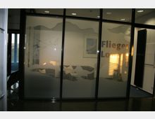 Eingangsbereich einer Lounge mit einer gläsernen Trennwand, die von einer mattierten Flächenbeklebung mit Wolken- und Flugzeugmotiven verziert ist. Der Schriftzug "Flieger Lounge" ist sichtbar, dahinter befindet sich ein beleuchteter Innenraum mit Sitzmöbeln.