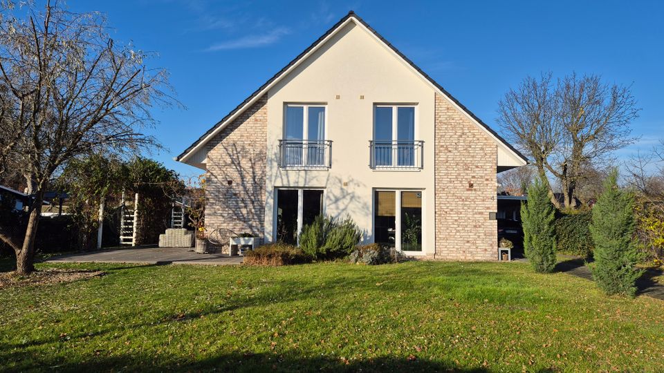 Zweistöckiges Einfamilienhaus mit Satteldach und heller Fassade, teilweise mit Backstein verkleidet. Im Vordergrund ein gepflegter Garten mit Rasen, Bäumen und einer Terrasse auf der linken Seite.