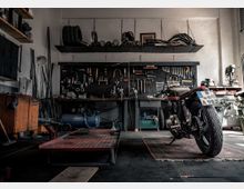 Werkstatt mit einem Motorrad im Vordergrund, auf einem Teppich geparkt. Im Hintergrund befinden sich eine Werkbank mit Werkzeugen an der Wand, Regale mit Reifen und Metallteilen sowie ein Kompressor.