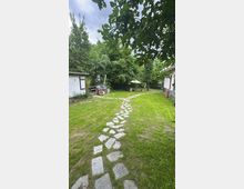 Garten mit grünem Rasen, geschmückt von einem gewundenen Weg aus unregelmäßigen Steinplatten. Umgeben von dichten Bäumen und Sträuchern, befindet sich links ein kleines Gebäude und rechts ein weiteres Gebäude mit hellem Anstrich.