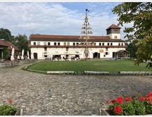 Historisches Gebäude mit weißer Fassade und roten Dachziegeln, im Vordergrund ein gepflasterter Platz und eine Rasenfläche. Ein zentraler Maibaum sowie offene Sitzbereiche mit Sonnenschirmen deuten auf eine Gastronomienutzung hin.