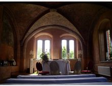 Innenansicht eines historischen Gewölberaumes mit bemalter Decke. Drei hohe Fenster mit Blick auf Bäume, ein mit weißen Stoffen gedeckter Altar, Stühle sowie Kerzenständer prägen den Raum.