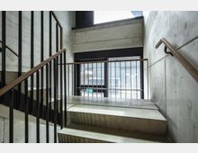 Eine moderne Betontreppe mit Metallgeländer und Holzhandlauf in einem Innenbereich. Große Fenster im Hintergrund lassen Tageslicht herein und geben den Blick auf eine Außenfassade frei.