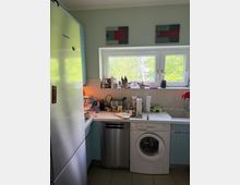 Kleine Küchenzeile mit weißer Arbeitsplatte unter einem breiten Doppelfenster, davor Spülbecken und verschiedene Küchenutensilien. Unter der Arbeitsplatte stehen eine Spülmaschine und eine Waschmaschine; links ein hoher Kühlschrank und hellblaue Schränke.