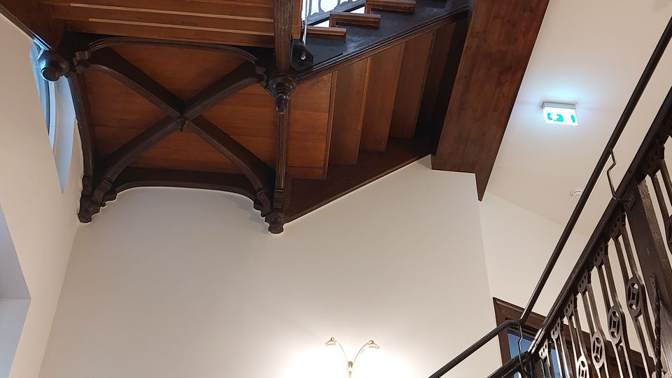 Holztreppenhaus mit verziertem Geländer und sichtbaren tragenden Holzbalken unter der Decke. Die helle Wand und eine stilvolle Wandlampe ergänzen die Zwischengeschosse, während ein Notausgangsschild in der oberen Ecke sichtbar ist.