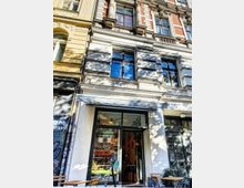 Fassade eines Altbaus mit reich verzierten Fenstern im oberen Bereich. Im Erdgeschoss befindet sich ein Café mit großer Glasfront, Markise, und Sitzgelegenheiten auf dem Bürgersteig.