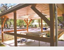 Holzüberdachte Grillstelle mit einem Metallgrill und Betonboden in einem Park. Im Hintergrund sind Bäume, Stehtische und weitere Elemente wie Schachspielmuster und ein Fahrzeug zu sehen.