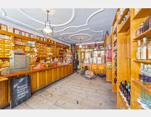 Innenraum eines historischen Ladens mit Holzverkleidung, einer Theke und Regalen voller Glasflaschen, Dosen und weiterer Produkte. Im Hintergrund sind ein gusseiserner Ofen, ein Waage und dekorative Elemente zu sehen, ergänzt durch eine verzierte Decke mit Stuckmustern.