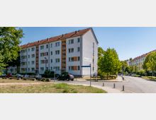 Mehrstöckiges Wohngebäude mit weißer Fassade und roten Dachziegeln, umgeben von Bäumen und einer Straße mit geparkten Autos. Im Vordergrund befindet sich ein kleiner Grünstreifen und ein Straßenschild, das auf die „Geschwister-Scholl-Straße“ hinweist.