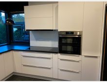 Moderne Küche mit weißen Schränken, schwarzer Arbeitsplatte und integriertem Elektroherd. Eine schwarze Hängelampe hängt über der Arbeitsfläche, und große Fenster bieten Blick ins Freie.