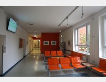 Wartebereich mit orangefarbenen Kunststoffstühlen und kleinen Tischen entlang einer Fensterfront. Der Raum hat weiße Wände, eine rote Akzentwand im Hintergrund, eine Deckenbeleuchtung und einen Flachbildschirm an der Wand.