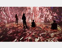 Ein dunkler Raum mit hellen, abstrakten Projektionen in Rot- und Orangetönen, die sowohl die Wände als auch den Boden bedecken. Vier Personen stehen oder sitzen in der Mitte des Raums und betrachten die Lichtinstallationen.