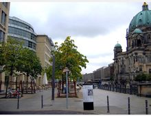Straßenszene in einer städtischen Umgebung mit einem modernen, abgerundeten Bürogebäude links, kleinen Bäumen und Sitzbereichen in der Mitte sowie dem Berliner Dom rechts im Bild. Im Hintergrund ist ein Fluss mit Geländern am Ufer sichtbar.