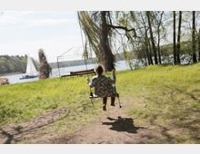 Ein Kind schaukelt auf einer Holzschaukel unter einem Baum in einer Wiese, mit Blick auf einen See im Hintergrund, auf dem ein Segelboot fährt. Im Hintergrund sind Bäume, eine Metallstruktur und eine Holzbank zu sehen.