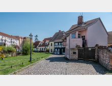 Kopfsteinpflasterstraße in einem historischen Wohnviertel mit kleinen Fachwerk- und farbigen Häusern, einer Laterne und angrenzenden Grünflächen sowie moderneren Gebäuden im Hintergrund.