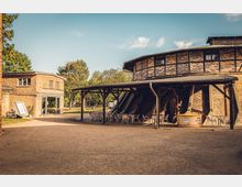Historisches Backsteingebäude in einem Außenbereich, bestehend aus einem runden Hauptbau mit Fachwerkelementen und einem Nebenbau. Unter einem überdachten Bereich stehen Stühle und Tische, umgeben von Bäumen und einer parkähnlichen Landschaft.