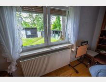 Blick aus einem kleinen Zimmer durch ein zweiflügeliges Fenster mit Spitzenvorhängen und Rollladen auf einen Garten mit Rasen, Schuppen und Zaun. Unter dem Fenster befindet sich ein Heizkörper; im Raum sind ein Stuhl und ein Regal zu sehen.