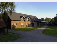 Ein ländlicher Hof mit einem großen, langgestreckten Gebäude aus Holz und Ziegeln mit einem dunkelgrauen Satteldach. Blumen schmücken die Fenster, und ein gepflasterter Weg führt durch die grüne Wiese, umgeben von weiteren kleineren Holzhütten und Bäumen im Hintergrund.
