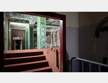 Blick durch einen Türrahmen in das Innere eines verlassenen Gebäudes mit rot gemusterten Stufen, abblätternder Deckenfarbe und grünen, teils mit Graffiti bedeckten Pfeilern. Im Hintergrund sind alte Holzverkleidungen, Fenster und stark verwitterte Wände zu sehen.