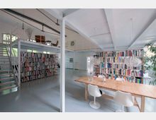 Großer, moderner Arbeitsraum in einem offenen Loft mit weiß gestrichenen Wänden und Decken. Im Vordergrund stehen ein langer Holztisch mit weißen Stühlen, im Hintergrund sind hohe Bücherregale, eine Treppe zur Galerieebene und große Fenster, die den Raum mit Tageslicht durchfluten.