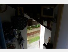 Blick aus einem dunklen, mit Bildern und Gegenständen dekorierten Raum durch eine geöffnete Tür auf einen kleinen Weg und grüne Pflanzen im Freien. Die Tür führt direkt ins Freie, und Licht fällt von außen herein.