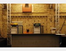 Industrie-ähnlicher Innenraum mit einer Bar im Vordergrund, dahinter zwei leere Getränkekühlschränke (mit den Marken "Beck's" und "Früh Kölsch Gold") vor einer gelben Backsteinwand mit Heizkörper und orangefarbener Lüftungsöffnung. Links und rechts befinden sich Metallträger, die Teil der Konstruktion sind.