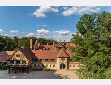 Großes historisches Fachwerkgebäude mit roten Ziegeldächern, zahlreichen Schornsteinen und einem Torbogen in der Mitte, umgeben von Bäumen und einem offenen Kiesplatz unter klarem Himmel mit vereinzelten Wolken.