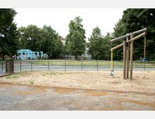 Spielplatz mit einem Holzgerüst und Schaukeln auf einer Sandfläche, im Hintergrund ein Park mit Bäumen, einer blauen Pergola und angrenzenden Gehwegen. Im weiteren Hintergrund sind Gebäude und einzelne Personen zu sehen.