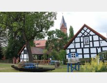 Fachwerkgebäude in ländlicher Umgebung mit weiß gestrichenen Wänden und schwarzen Holzstreben, umgeben von Bäumen und einem Vorgarten mit Dekorationen wie einer Holzbank und Milchkannen. Im Hintergrund ist ein Kirchturm aus rotem Backstein sichtbar.