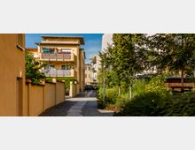 Gepflasterter Weg in einem Wohnviertel mit mehrstöckigen, gelb-beigen Gebäuden, Balkonen und umliegender Grünfläche. Bäume und Sträucher säumen den Weg, während im Hintergrund parkende Autos sichtbar sind.