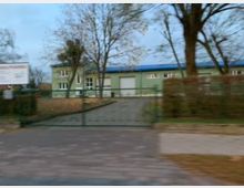 Ein eingeschossiges Gebäude mit grün gestrichener Fassade und blauer Dachverkleidung, umgeben von Bäumen und einem Zaun. Im Vordergrund ist eine geschlossene Einfahrt mit einem Metalltor zu sehen, daneben ein gepflasterter Gehweg.