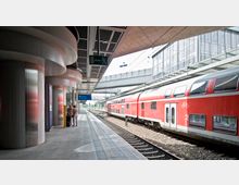 Ein moderner Bahnhof mit überdachtem Bahnsteig, einer roten Doppelstockbahn an den Gleisen und Personen, die auf dem Bahnsteig warten. Im Hintergrund sind Glasfassaden und eine Brücke sichtbar.