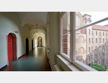 Ein langer, heller Flur mit gewölbter Decke, Hängelampen und roten Türen auf der linken Seite; rechts befinden sich große Fenster mit Blick auf ein Backsteingebäude im klassischen Stil. Draußen sind grüne Büsche und Architektur mit hohen, schmalen Fenstern sichtbar.