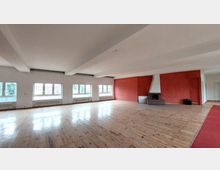 Großer, lichtdurchfluteter Raum mit Holzboden, großen Fenstern auf der linken Seite und einer roten Wand mit einem modernen Kamin auf der rechten Seite. Die Decke ist weiß mit sichtbaren Balken, und Heizkörper befinden sich unter den Fenstern.