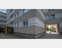 Mehrstöckiges Wohngebäude mit grauer Fassade und gelben Akzenten, darunter ein Eingangsbereich mit Glastür und Fahrradständern. Ein Durchgang führt zu einem begrünten Innenhof, eine Person mit Rollwagen ist im Bild zu sehen.