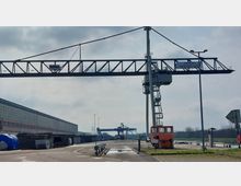 Industriehafen mit großem Portalkran, gekennzeichnet mit „Stadthafen Brandenburg a. d. H.“, einer angrenzenden Lagerhalle und gestapelten Materialien. Im Hintergrund sind weitere Kräne sowie ein Wasserweg sichtbar.