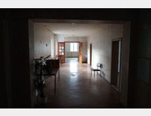 Dunkler Flur in einem Gebäude mit schlicht gestrichenen Wänden und Holzfußboden, der zu einem helleren Raum mit großen Fenstern und offenen Holztüren führt; links ein kleiner Tisch mit einer Pflanze, rechts ein einzelner Stuhl an der Wand.