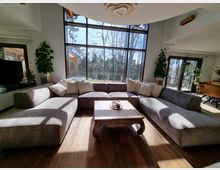 Modernes Wohnzimmer mit einem großen, grauen U-förmigen Sofa und einem Holztisch in der Mitte. Der Raum verfügt über hohe Fenster mit Blick auf eine Terrasse und einen bewaldeten Außenbereich.