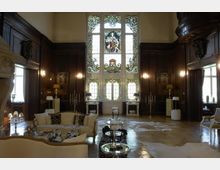 Ein eleganter Innenraum mit dunklen Holzvertäfelungen, hohen Decken und einem großen, verzierten Buntglasfenster als zentralem Element. Der Raum ist mit klassischen Möbeln, Kronleuchtern und Kerzenhaltern ausgestattet und vermittelt eine luxuriöse, historische Atmosphäre.