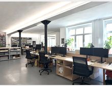 Heller, offener Büroraum im Loftstil mit langen Arbeitstischen, mehreren Monitoren und schwarzen Drehstühlen. Große Fensterfront mit Pflanzen auf der Fensterbank sowie offene Regale und Stauraumsysteme im Hintergrund; schwarze Stützsäulen tragen die Decke.