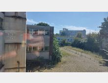 Blick auf ein vernachlässigtes Außengelände mit überwucherten Pflasterflächen und einem alten, verwitterten Gebäude mit mehreren Fenstern im Vordergrund. Im Hintergrund sind weitere Gebäude, Büsche und Bäume sichtbar, unter einem sonnigen, blauen Himmel.