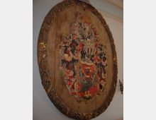 Holztafel mit einem abgeblätterten, bemalten Wappen, das filigrane Ornamente und ein schwarzes Tiermotiv auf rotem Hintergrund zeigt, umgeben von einem ovalen Rahmen mit floralen Schnitzereien. Die Tafel ist an einer weißen Wand angebracht, im Hintergrund ragt ein Teil eines Kronleuchters ins Bild.