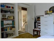 Ein Schlafzimmer mit weißen Wänden und Teppichboden in hellbrauner Farbe. Links steht ein Regal mit Büchern und Ordnern, rechts ein weißer Schrank und ein Holzstuhl. Im Hintergrund führt eine offene Tür zu einem Flur mit Treppe.