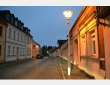 Straßenszene in einer Kleinstadt bei Dämmerung mit gepflasterter Fahrbahn, links und rechts stehen Häuser im traditionellen Baustil. Straßenlaternen werfen warmes Licht auf die Gebäude und den Gehweg, im Hintergrund sind Bäume und weitere Gebäude zu sehen.