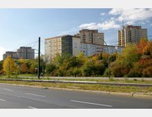 Straßenszene mit mehrspuriger Fahrbahn im Vordergrund, dahinter eine Straßenbahntrasse. Im Hintergrund stehen mehrere Hochhäuser, umgeben von herbstlich gefärbten Bäumen und Sträuchern.