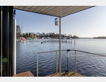 Blick von einer Terrasse mit Überdachung und Geländer auf einen See mit Stegen und Segelbooten. Im Hintergrund sind Bäume und Gebäude am Ufer zu sehen, bei klarem Himmel und Sonnenschein.