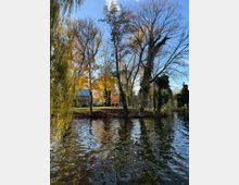 Ufer eines ruhigen Teichs mit spiegelndem Wasser, dahinter eine Grünfläche mit hohen Laub- und Weidenbäumen in herbstlichen Farben. Links steht ein kleines Gebäude mit hellem Giebel und Metalldach zwischen den Bäumen.