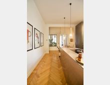 Moderne Küche mit hellem Fischgrät-Holzboden, einer Kücheninsel aus Edelstahl und hängenden Pendelleuchten. An der linken Wand hängen gerahmte Poster, und im Hintergrund befindet sich ein Fenster mit Blick auf Pflanzen.